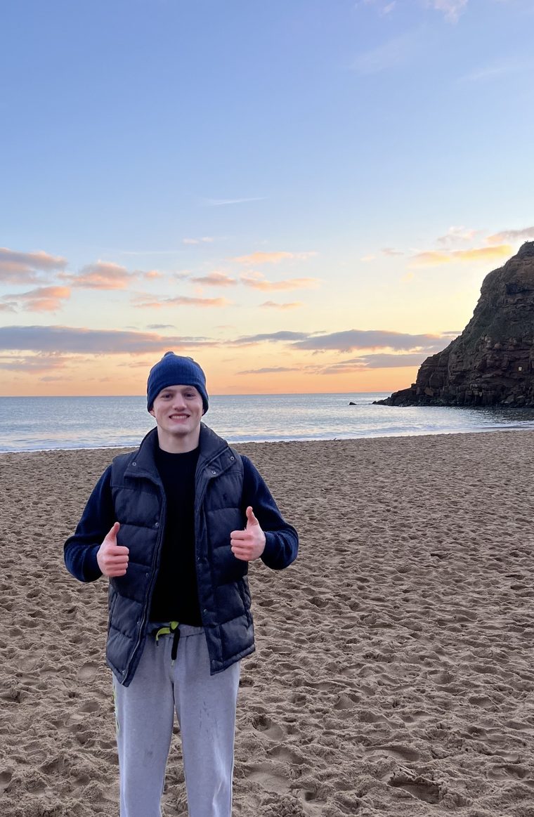 Joe on a beach at sunrise, wearing a beanie and giving two thumbs up.